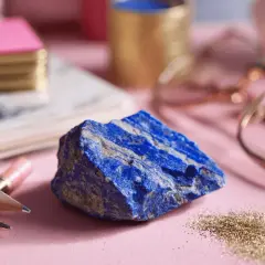 A raw, vibrant blue lapis lazuli stone sits on a pink surface, surrounded by gold pencils, a pink notebook, and rose gold glasses.