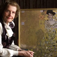 An elderly woman poses beside a framed woman in gold painting, her expression serene.