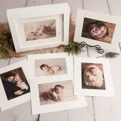 A collection of fine art photography prints showcasing a newborn baby and family. The images are displayed on a rustic wooden surface, with delicate greenery and burlap accents.