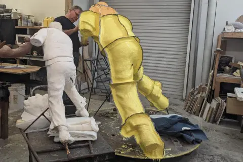Artist works on a yellow, textured sculpture in a studio, with a white plaster figure nearby. This casting in art is a complex process.