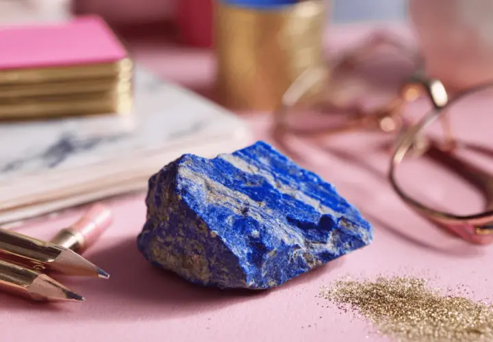 A raw, vibrant blue lapis lazuli stone sits on a pink surface, surrounded by gold pencils, a pink notebook, and rose gold glasses.