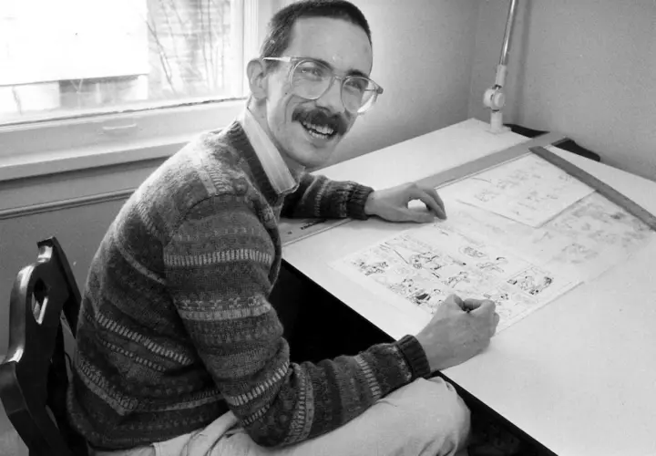 A smiling man with glasses and a mustache, likely Thomas Hobbes, draws a comic strip at his drafting table.