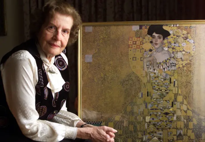 An elderly woman poses beside a framed woman in gold painting, her expression serene.