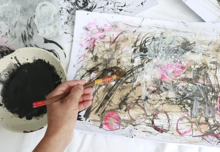 A hand holds a brush, adding bold strokes to an abstract drawing. Black ink splatters a palette beside the energetic artwork.