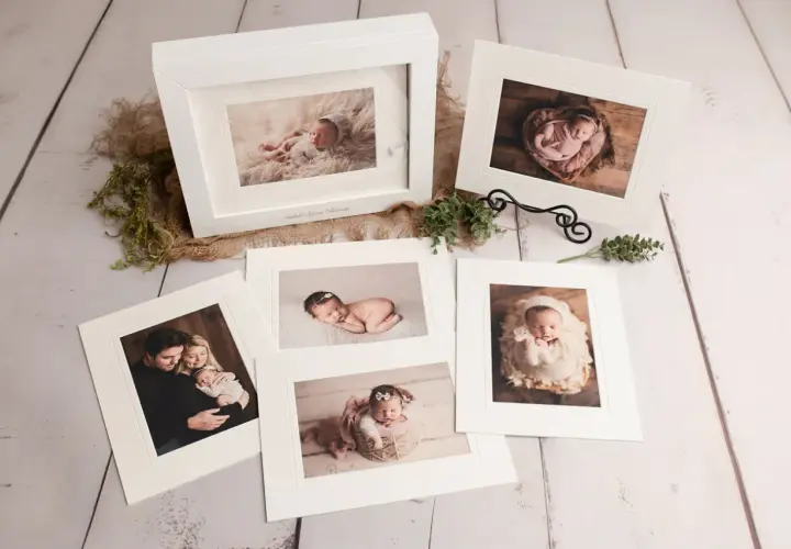 A collection of fine art photography prints showcasing a newborn baby and family. The images are displayed on a rustic wooden surface, with delicate greenery and burlap accents.