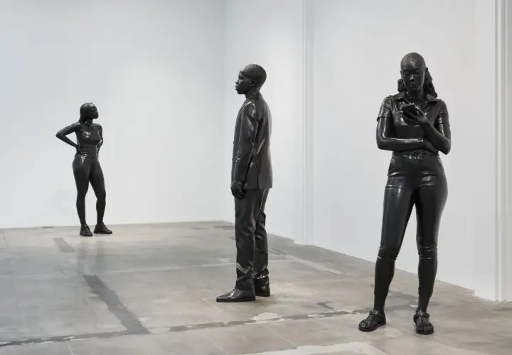 Three striking figures by thomas j price sculpture stand in a minimalist gallery space. One woman poses confidently, another is engrossed in her phone, and a man in a suit stands stoically.