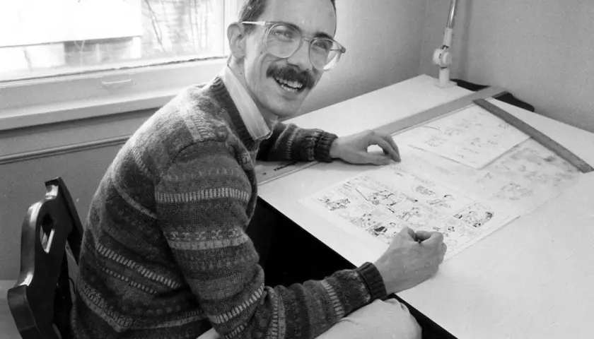A smiling man with glasses and a mustache, likely Thomas Hobbes, draws a comic strip at his drafting table.