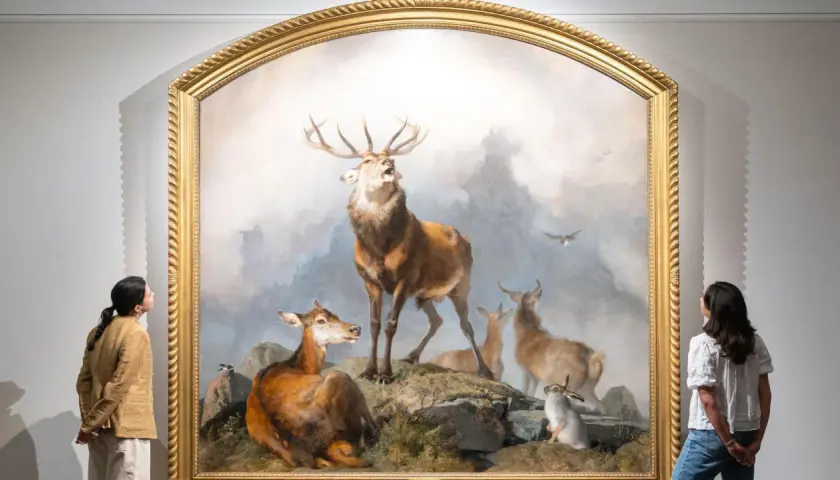 Two women admire a dramatic painting of a stag, deer, and rabbits, reminiscent of Edwin Landseer paintings, in a gallery.