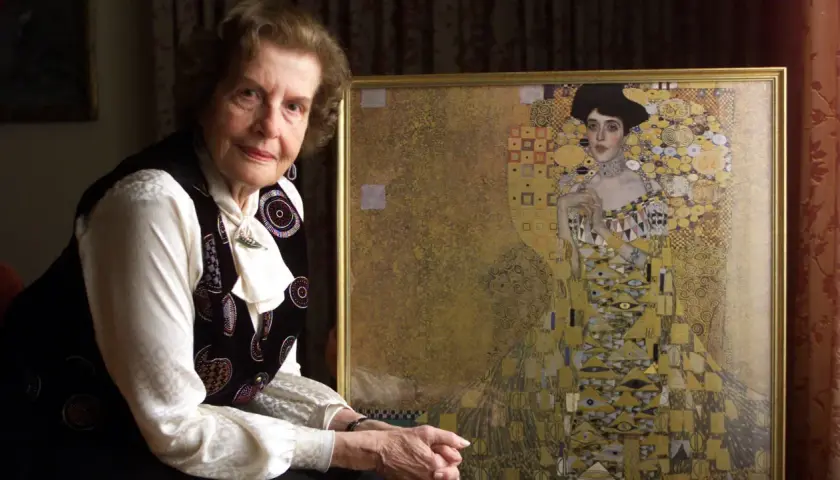 An elderly woman poses beside a framed woman in gold painting, her expression serene.