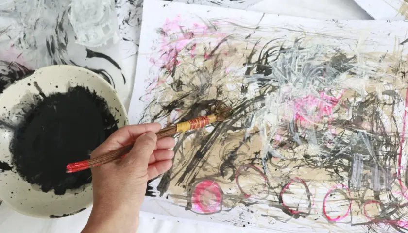 A hand holds a brush, adding bold strokes to an abstract drawing. Black ink splatters a palette beside the energetic artwork.