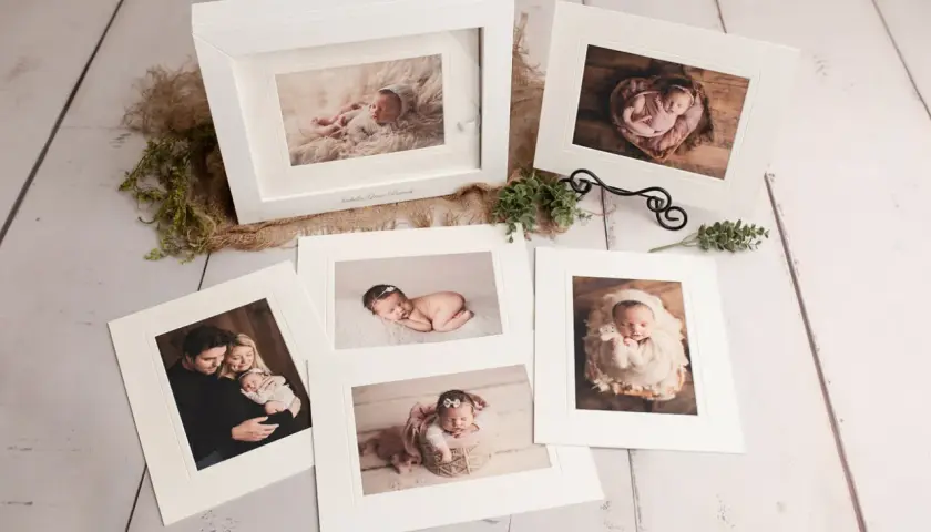 A collection of fine art photography prints showcasing a newborn baby and family. The images are displayed on a rustic wooden surface, with delicate greenery and burlap accents.