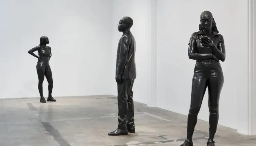 Three striking figures by thomas j price sculpture stand in a minimalist gallery space. One woman poses confidently, another is engrossed in her phone, and a man in a suit stands stoically.