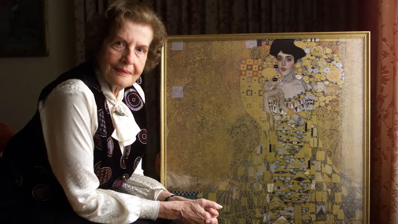 An elderly woman poses beside a framed woman in gold painting, her expression serene.
