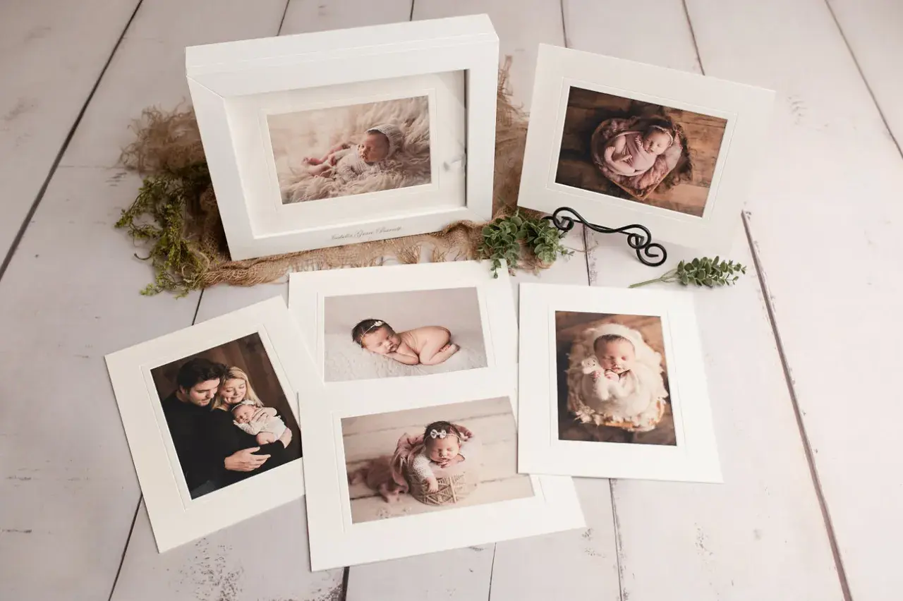 A collection of fine art photography prints showcasing a newborn baby and family. The images are displayed on a rustic wooden surface, with delicate greenery and burlap accents.