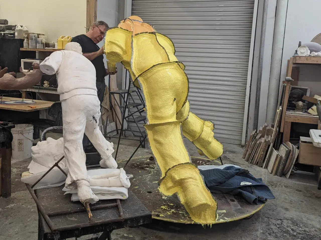 Artist works on a yellow, textured sculpture in a studio, with a white plaster figure nearby. This casting in art is a complex process.