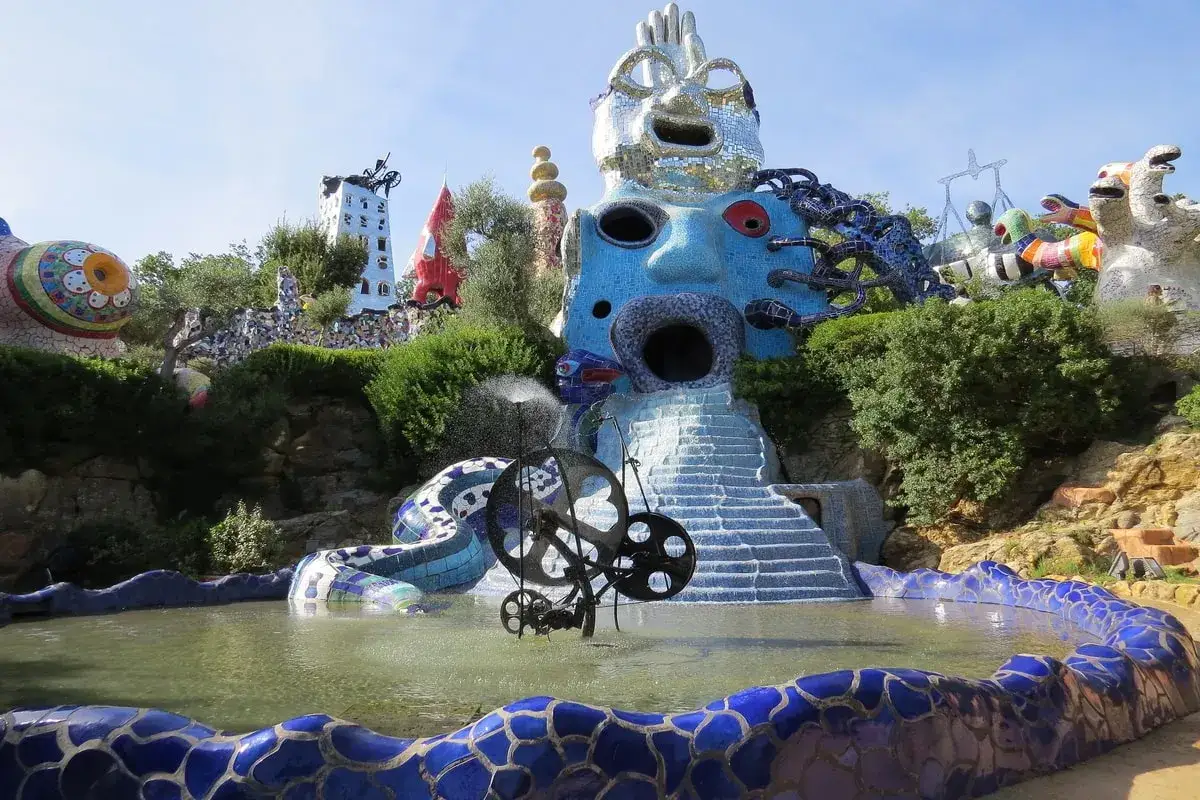 A vibrant, fantastical landscape featuring Niki de Saint Phalle's Nanas sculptures, including a giant blue face fountain and a mosaic snake.