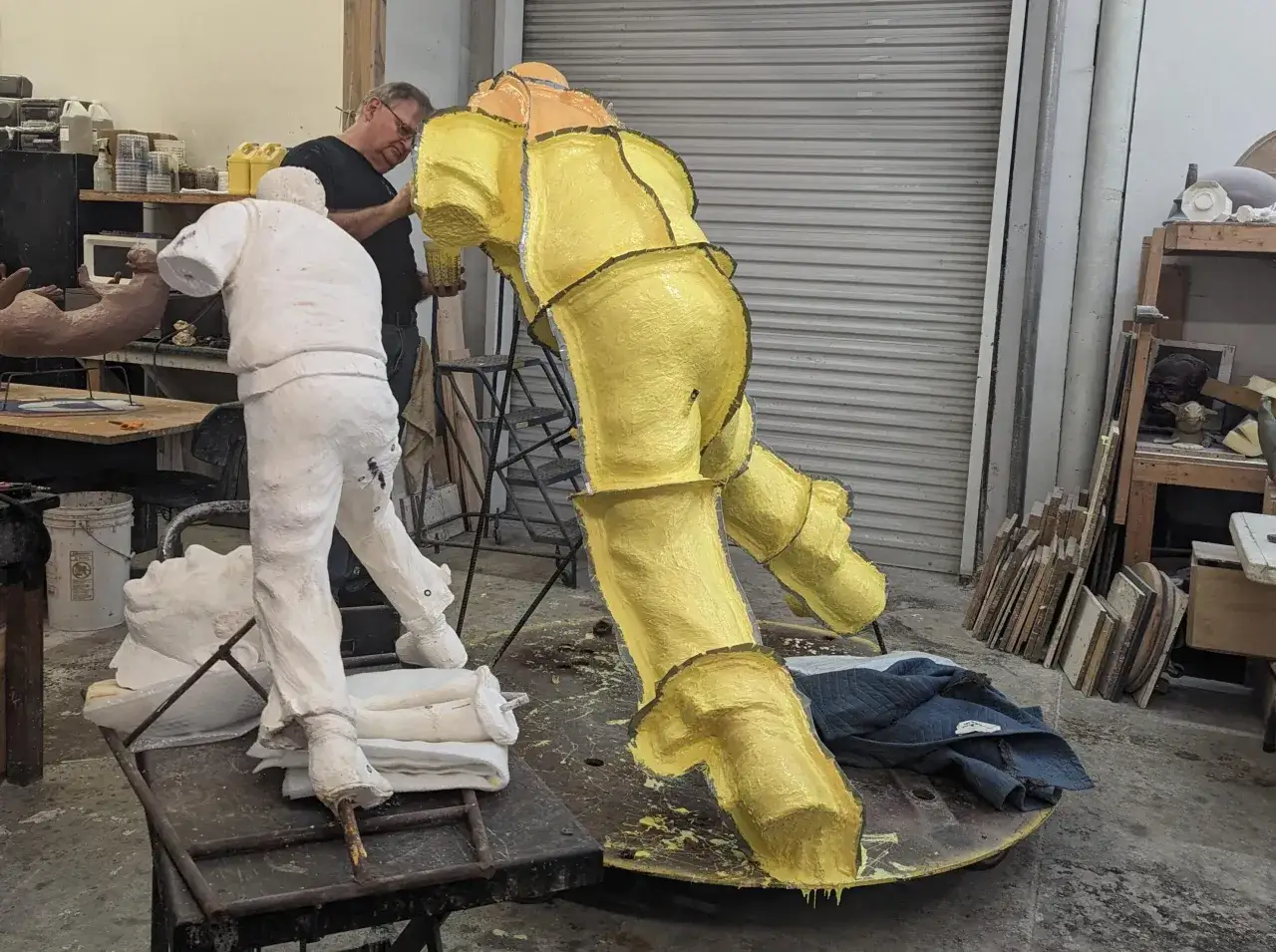 Artist works on a yellow sculpture, a white figure nearby. The process of casting art is underway in a studio.
