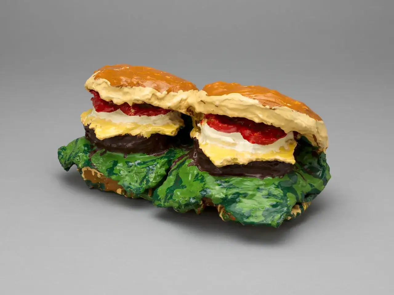 Two colorful, textured sculptures of hamburgers, reminiscent of Andy Warhol's iconic pop art, sit side-by-side on a gray background.