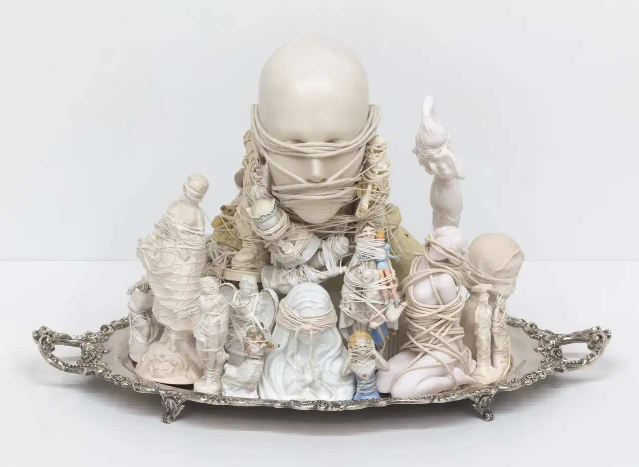 A surreal found object art piece on a silver tray, featuring a large doll head and various ceramic figures bound with rope.