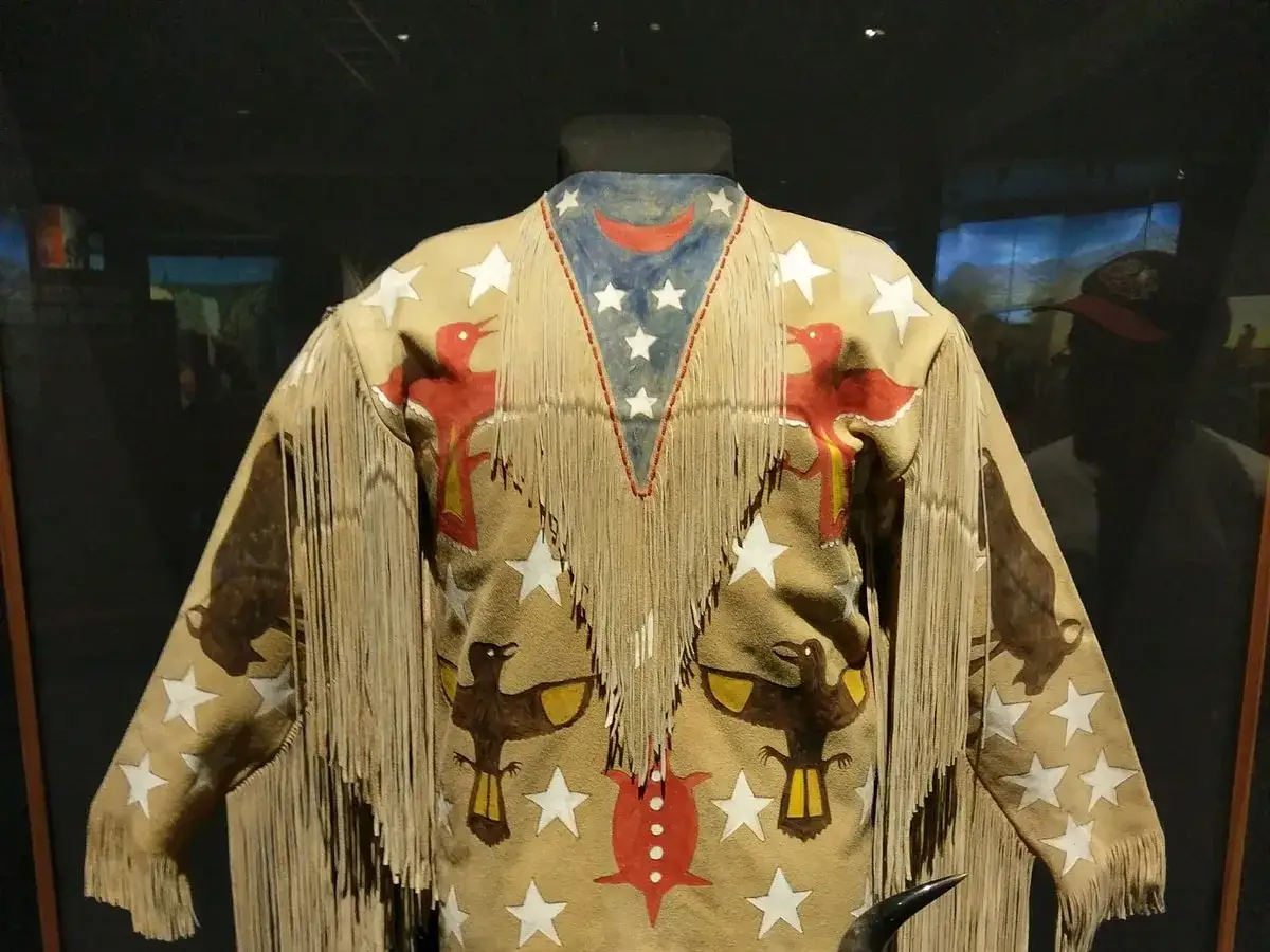 A fringed hide shirt adorned with painted ghost dance symbols: red birds, buffalo, and stars.