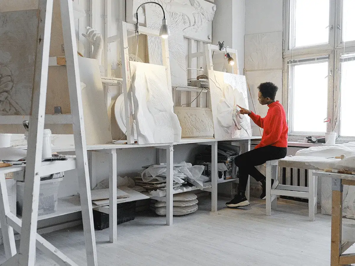 An artist in a red sweater examines a textured relief, a glimpse into the creation of fine art in a bright, white studio filled with sculptures and materials.