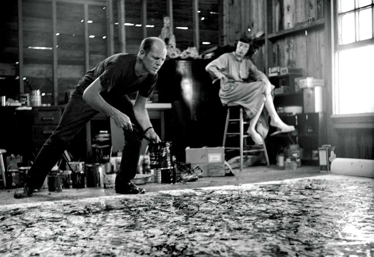Jackson Pollock's intense Jackson Pollock face is captured as he drips paint onto a canvas. A woman watches from a stool in his studio.