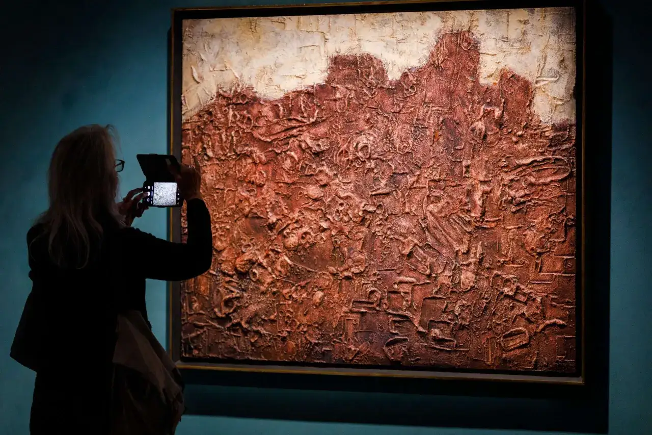 A visitor photographs a textured, abstract painting in the style of Jean Dubuffet art, featuring earthy tones and a rough, impasto surface.