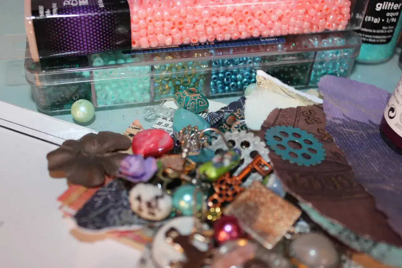 A collage of craft supplies: beads in pink, teal, and blue, charms, gears, and torn paper pieces.
